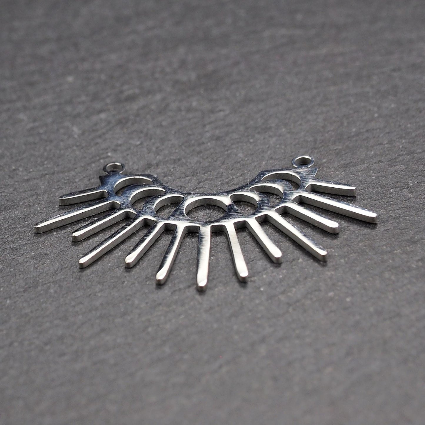 Versilberter Metallverbinder, halbkreisförmig und durchbrochen, mit rundem Mittelloch, mehreren schmalen radialen Strahlen, glänzender Oberfläche und zwei kleinen Ösen oben.