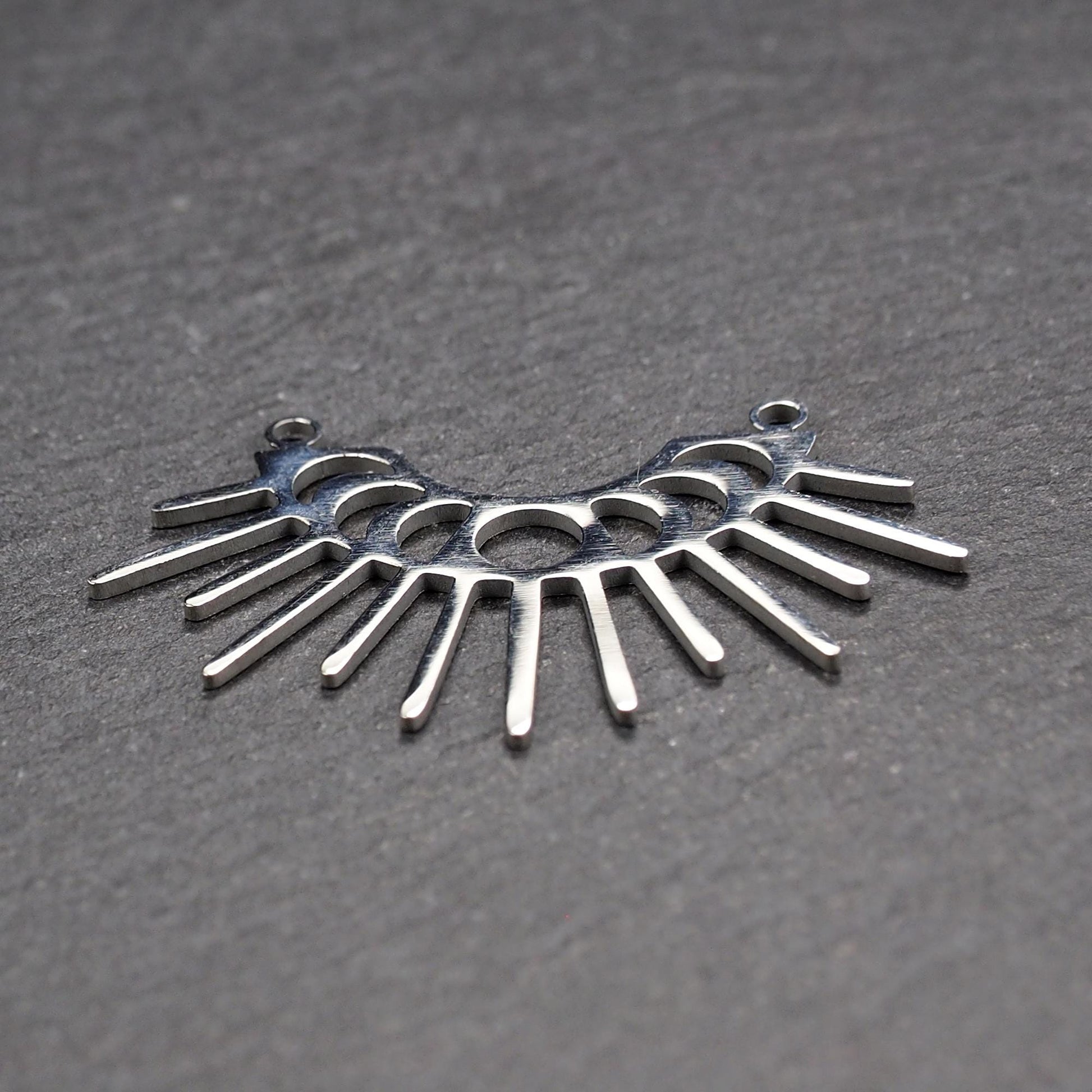 Versilberter Metallverbinder, halbkreisförmig und durchbrochen, mit rundem Mittelloch, mehreren schmalen radialen Strahlen, glänzender Oberfläche und zwei kleinen Ösen oben.