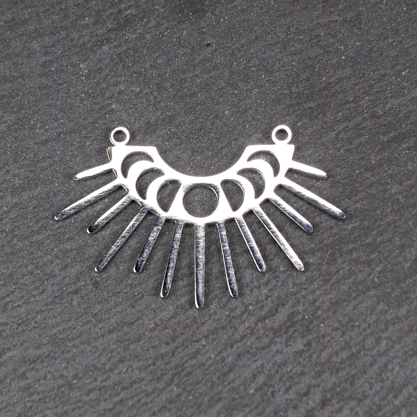 Versilberter Metallverbinder, halbkreisförmig und durchbrochen, mit rundem Mittelloch, mehreren schmalen radialen Strahlen, glänzender Oberfläche und zwei kleinen Ösen oben.
