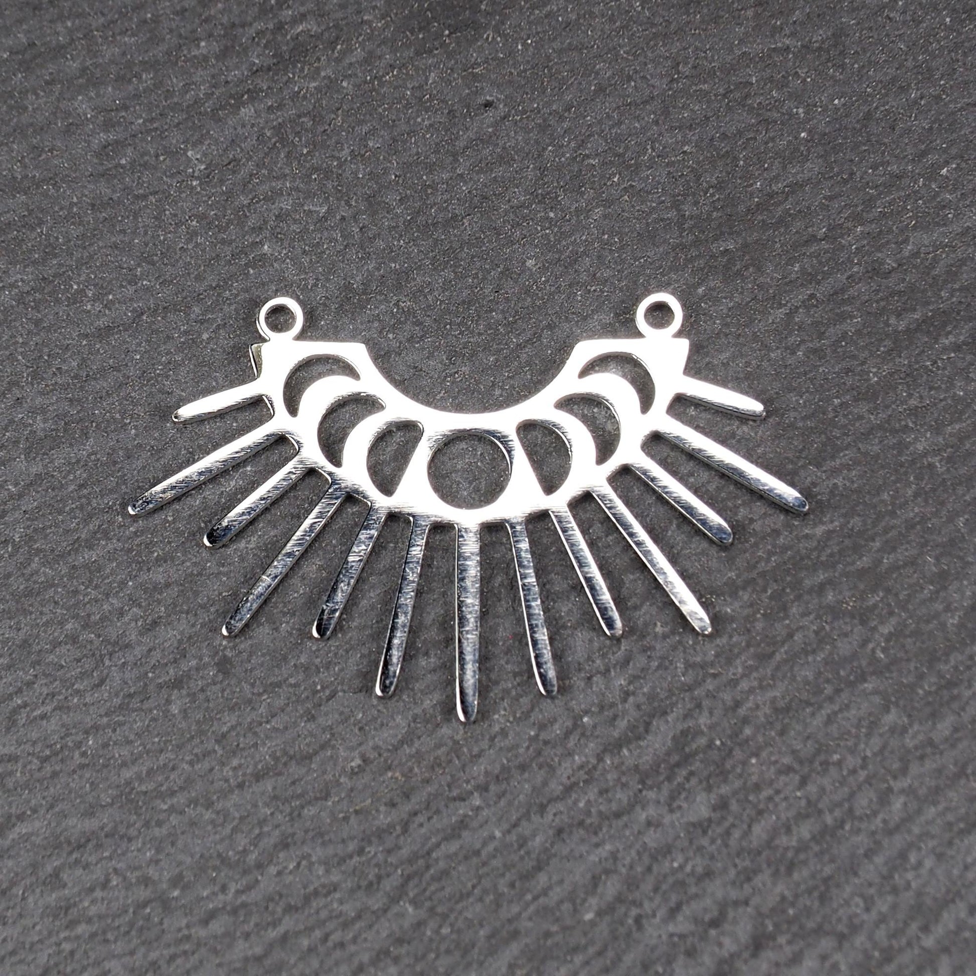 Versilberter Metallverbinder, halbkreisförmig und durchbrochen, mit rundem Mittelloch, mehreren schmalen radialen Strahlen, glänzender Oberfläche und zwei kleinen Ösen oben.