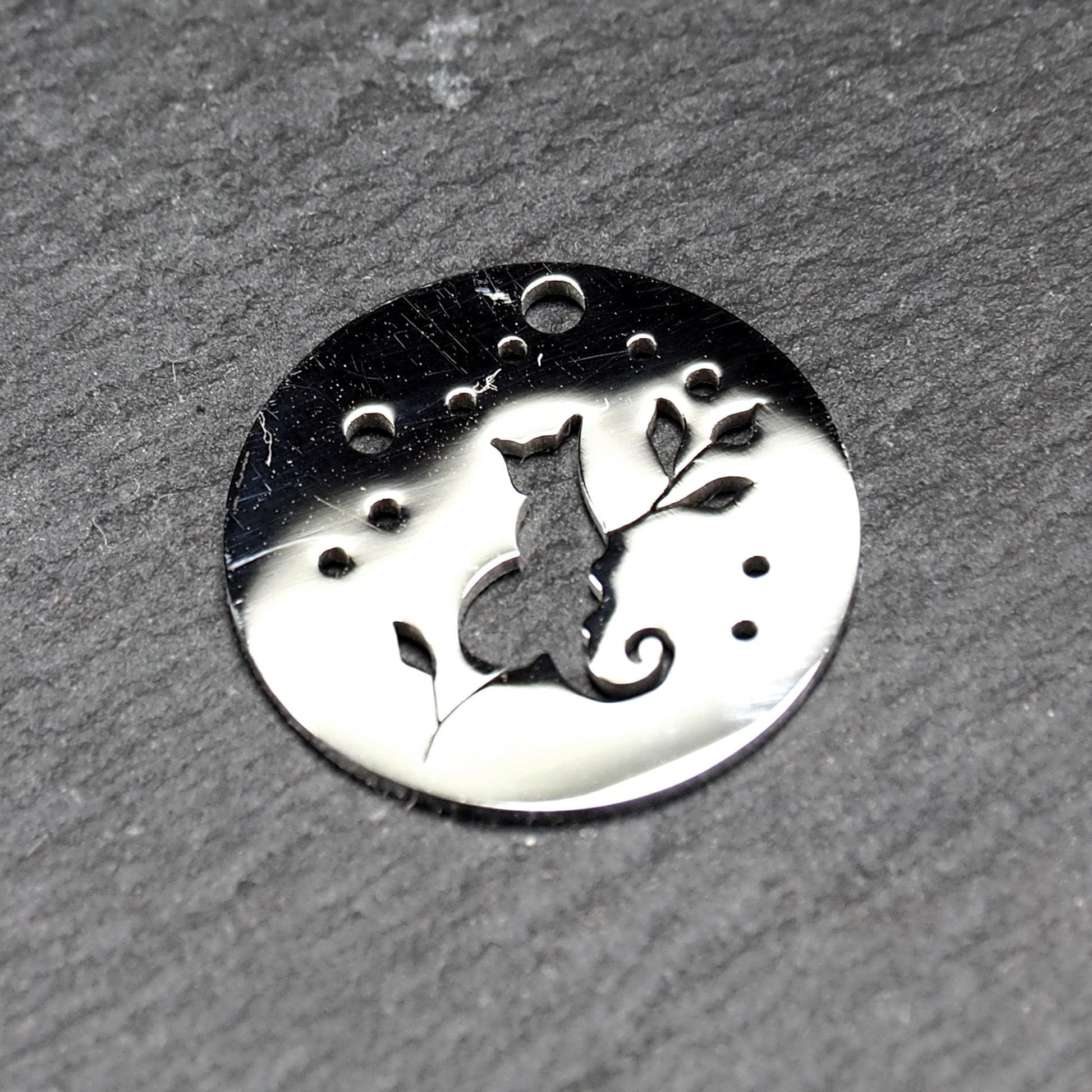 Runder, flacher Anhänger aus poliertem Edelstahl, Cut-out mit sitzender Katzen-Silhouette und Blattzweig, Öse oben und mehrere kleine Bohrungen.
