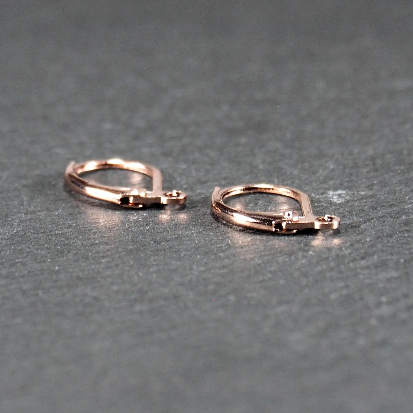 Zwei rosévergoldete Klappbrisuren in glänzender Metalloptik, ovale Leverback-Form mit sichtbarer kleinen Öse am unteren Ende, auf dunklem Hintergrund.