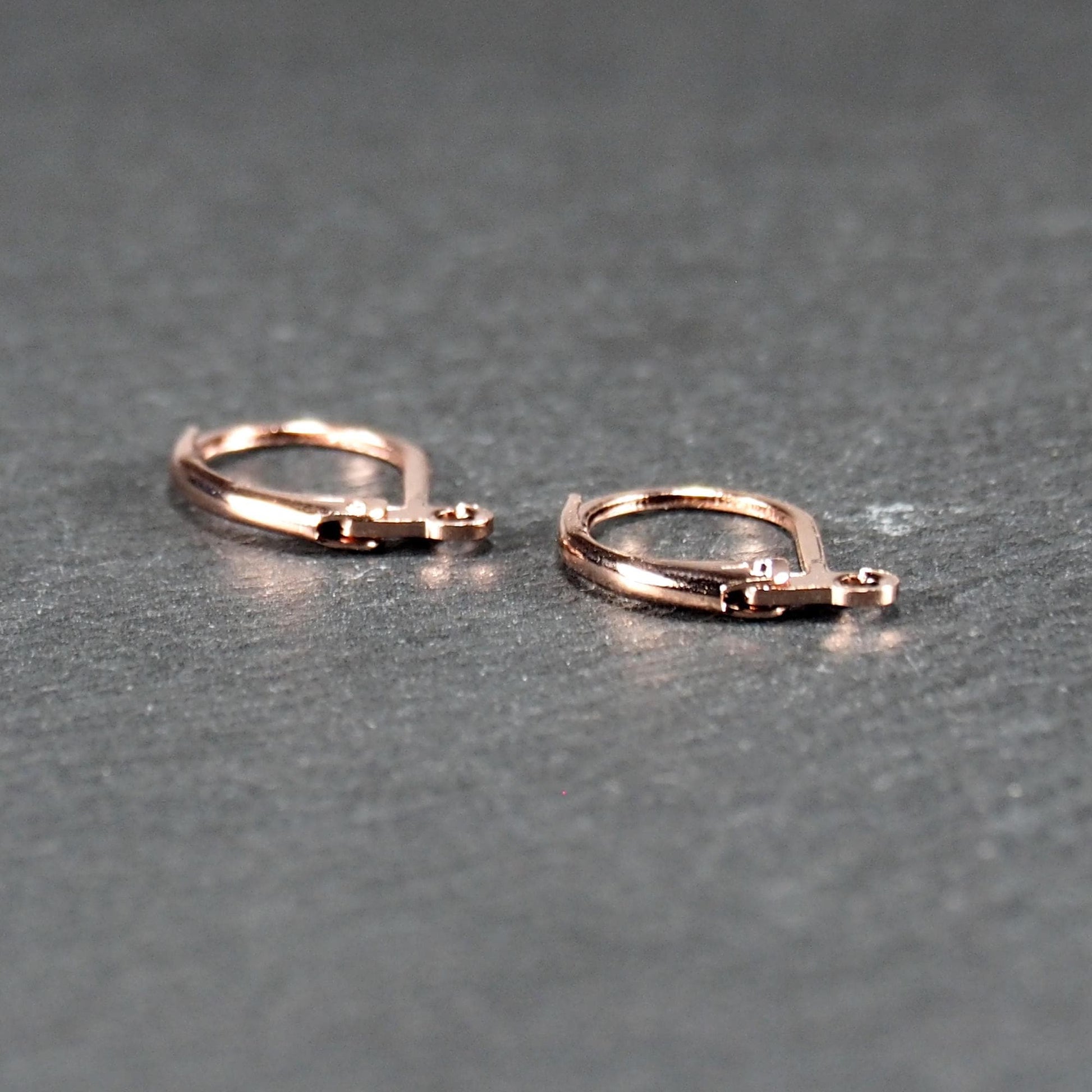 Zwei rosévergoldete Klappbrisuren in glänzender Metalloptik, ovale Leverback-Form mit sichtbarer kleinen Öse am unteren Ende, auf dunklem Hintergrund.
