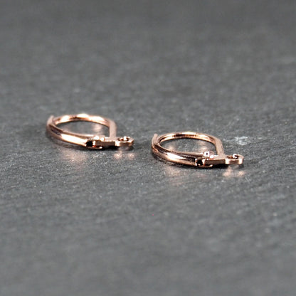 Zwei rosévergoldete Klappbrisuren in glänzender Metalloptik, ovale Leverback-Form mit sichtbarer kleinen Öse am unteren Ende, auf dunklem Hintergrund.