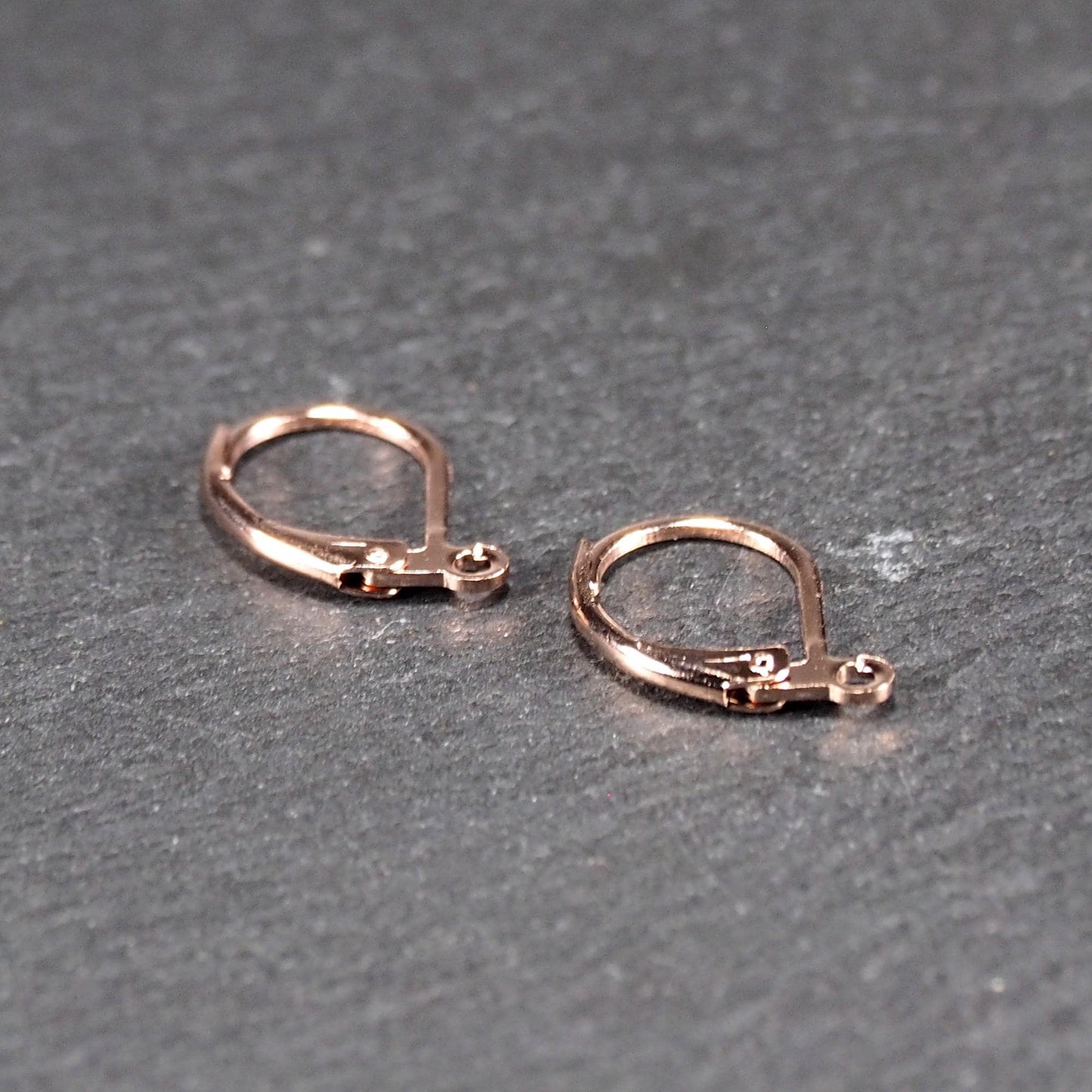 Zwei rosévergoldete Klappbrisuren in glänzender Metalloptik, ovale Leverback-Form mit sichtbarer kleinen Öse am unteren Ende, auf dunklem Hintergrund.