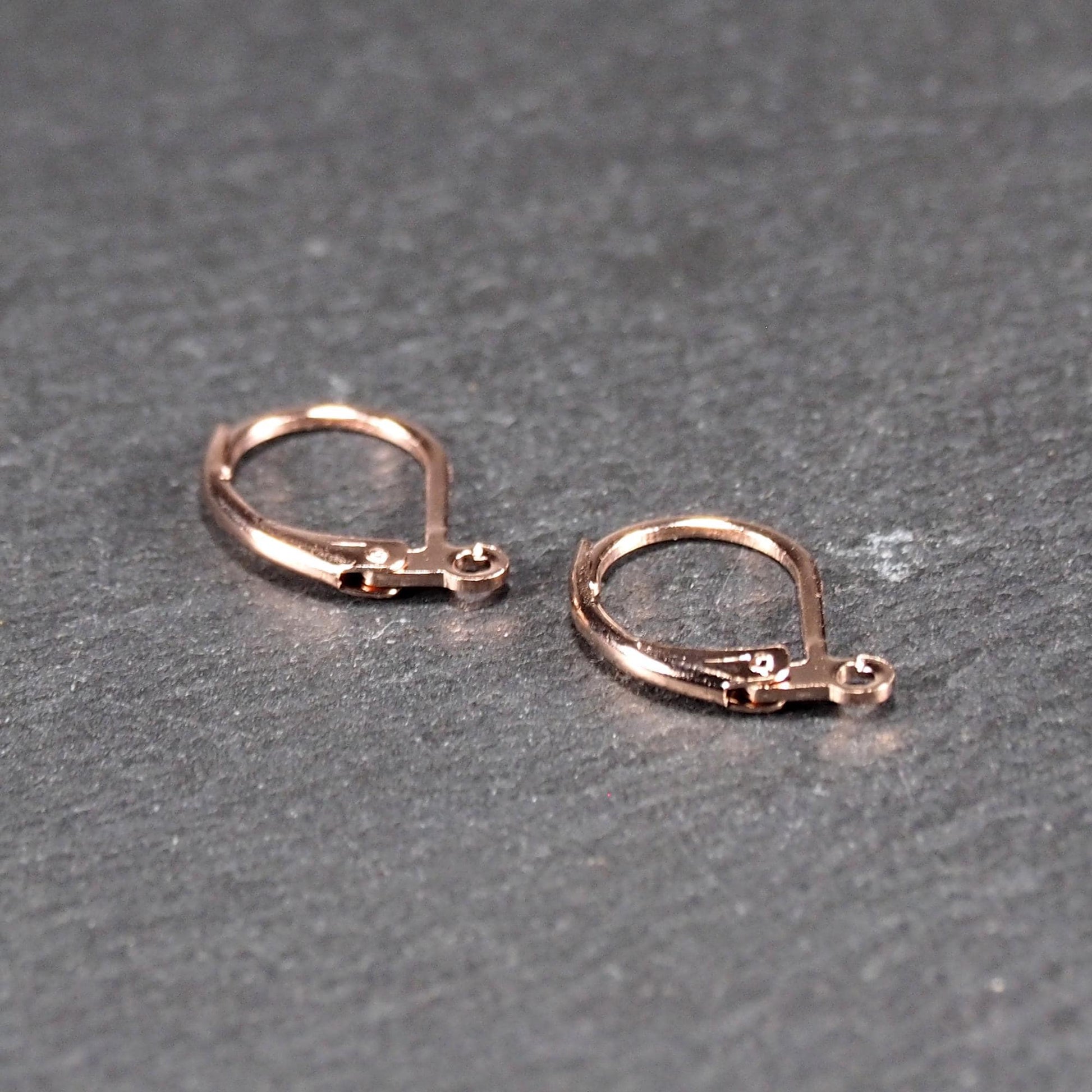 Zwei rosévergoldete Klappbrisuren in glänzender Metalloptik, ovale Leverback-Form mit sichtbarer kleinen Öse am unteren Ende, auf dunklem Hintergrund.