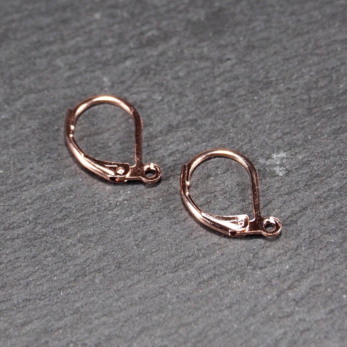 Zwei rosévergoldete Klappbrisuren in glänzender Metalloptik, ovale Leverback-Form mit sichtbarer kleinen Öse am unteren Ende, auf dunklem Hintergrund.