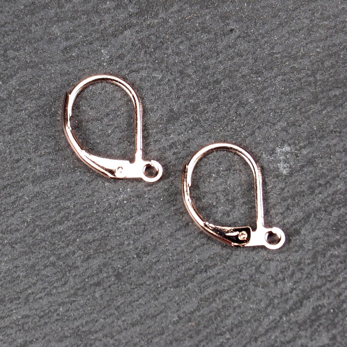 Zwei rosévergoldete Klappbrisuren in glänzender Metalloptik, ovale Leverback-Form mit sichtbarer kleinen Öse am unteren Ende, auf dunklem Hintergrund.