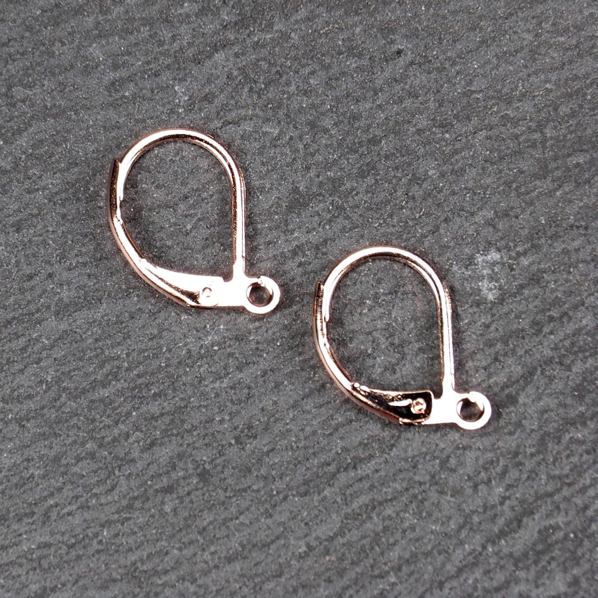 Zwei rosévergoldete Klappbrisuren in glänzender Metalloptik, ovale Leverback-Form mit sichtbarer kleinen Öse am unteren Ende, auf dunklem Hintergrund.