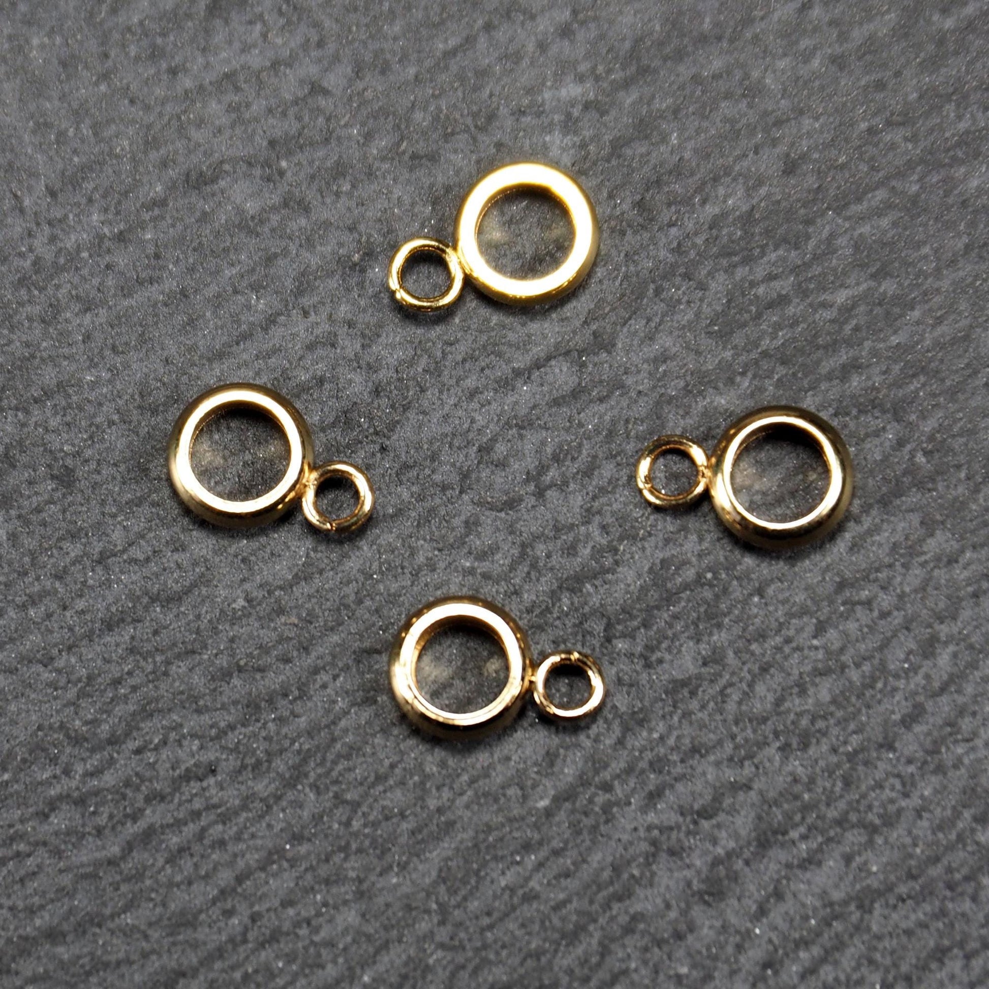 Vier vergoldete ringförmige Anhängerösen mit glänzender goldfarbener Oberfläche, hohlem Mittelring und jeweils kleinem seitlichem Ösenring.