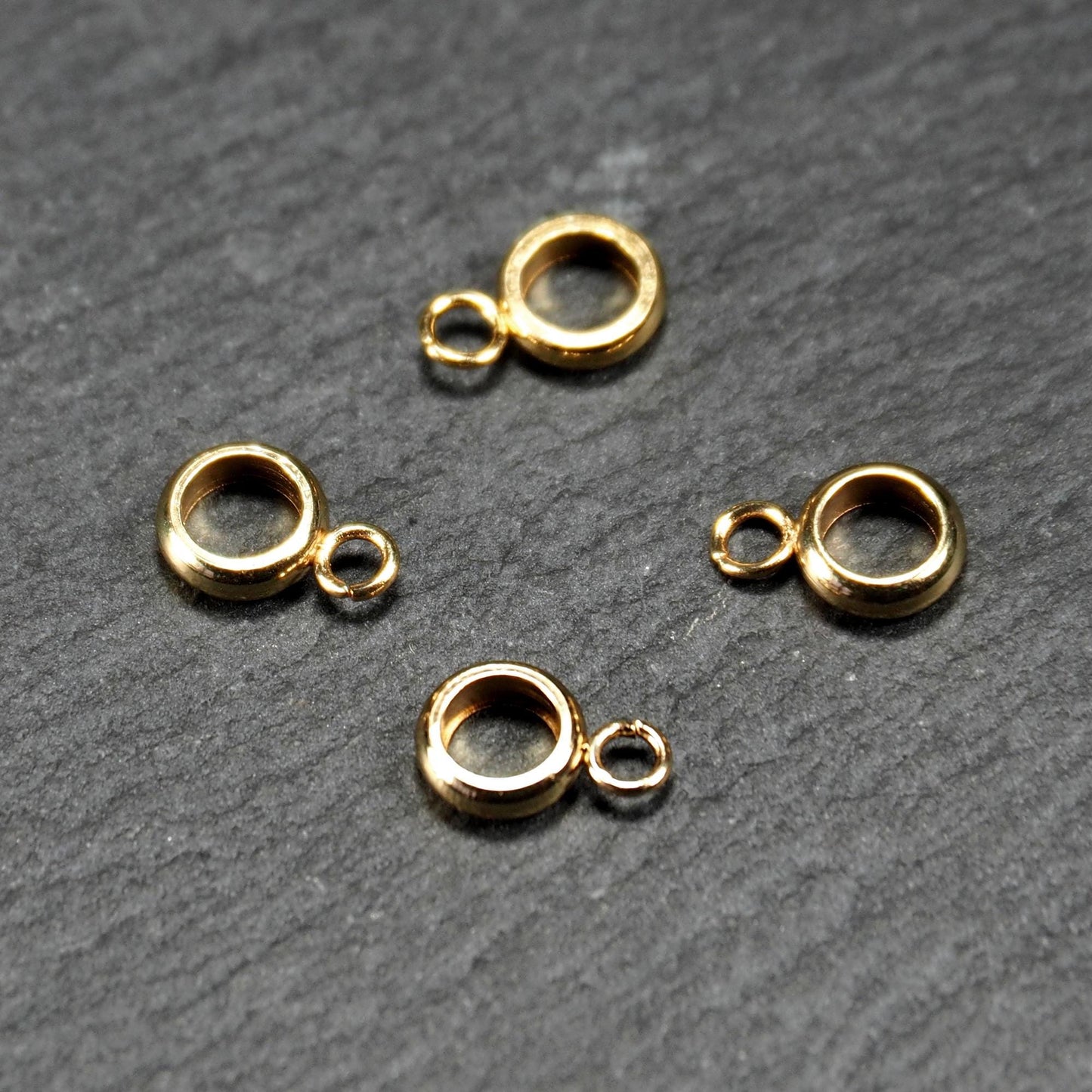 Vier vergoldete ringförmige Anhängerösen mit glänzender goldfarbener Oberfläche, hohlem Mittelring und jeweils kleinem seitlichem Ösenring.