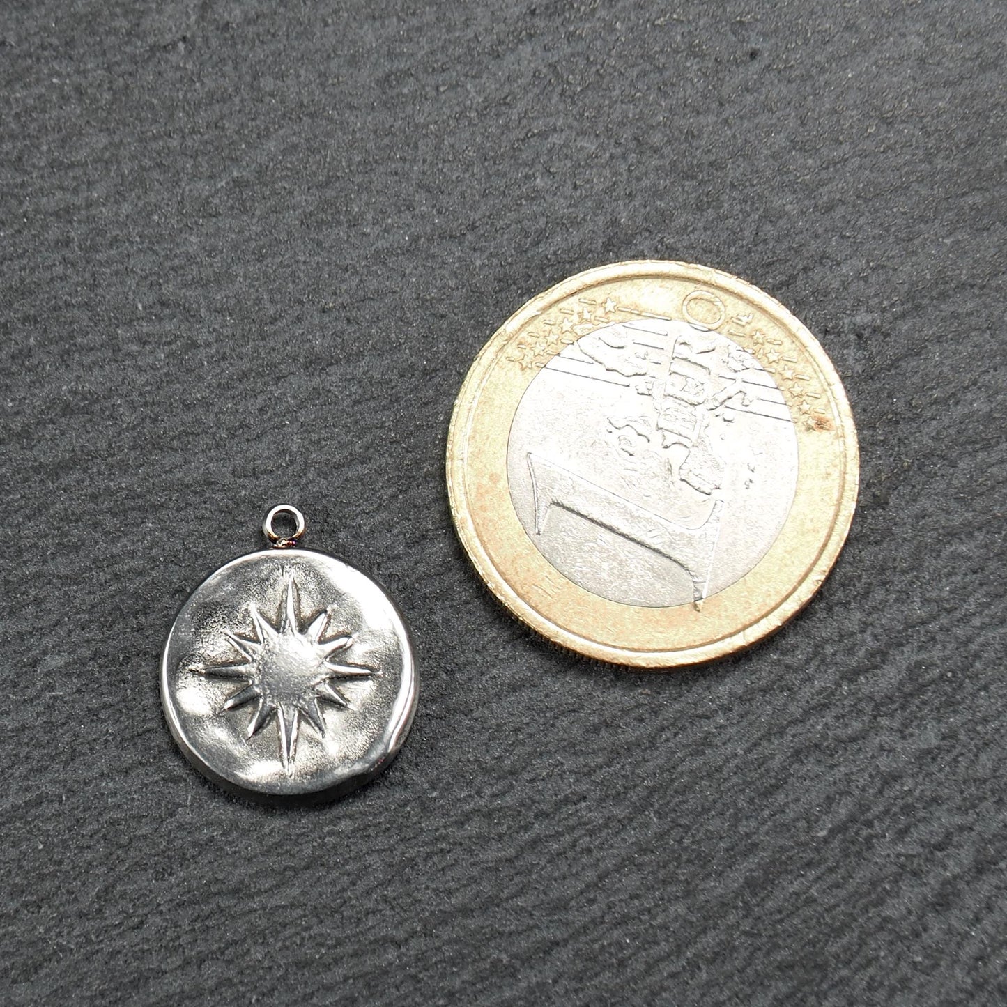 Runder silberner Metallanhänger mit reliefartigem Sonnen- und Kompassmotiv, mit Öse oben; neben einer Ein-Euro-Münze zur Größendarstellung.