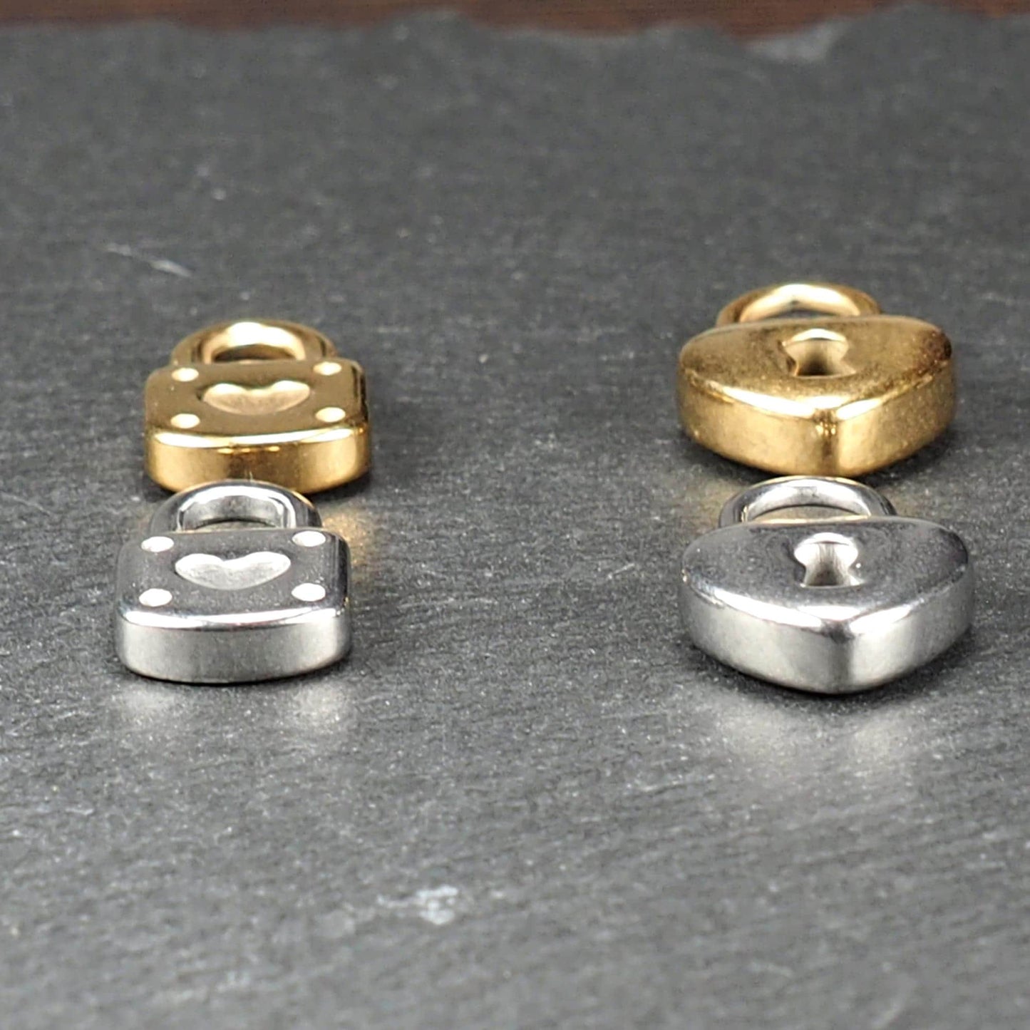 Vier glänzende Metallanhänger auf dunklem Untergrund: zwei vergoldete und zwei silberne Schloss- und Herz-Schlüssel-Motiv-Anhänger mit sichtbarer Öse.