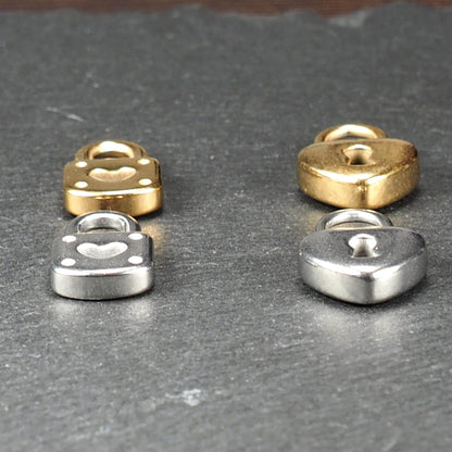 Vier glänzende Metallanhänger auf dunklem Untergrund: zwei vergoldete und zwei silberne Schloss- und Herz-Schlüssel-Motiv-Anhänger mit sichtbarer Öse.