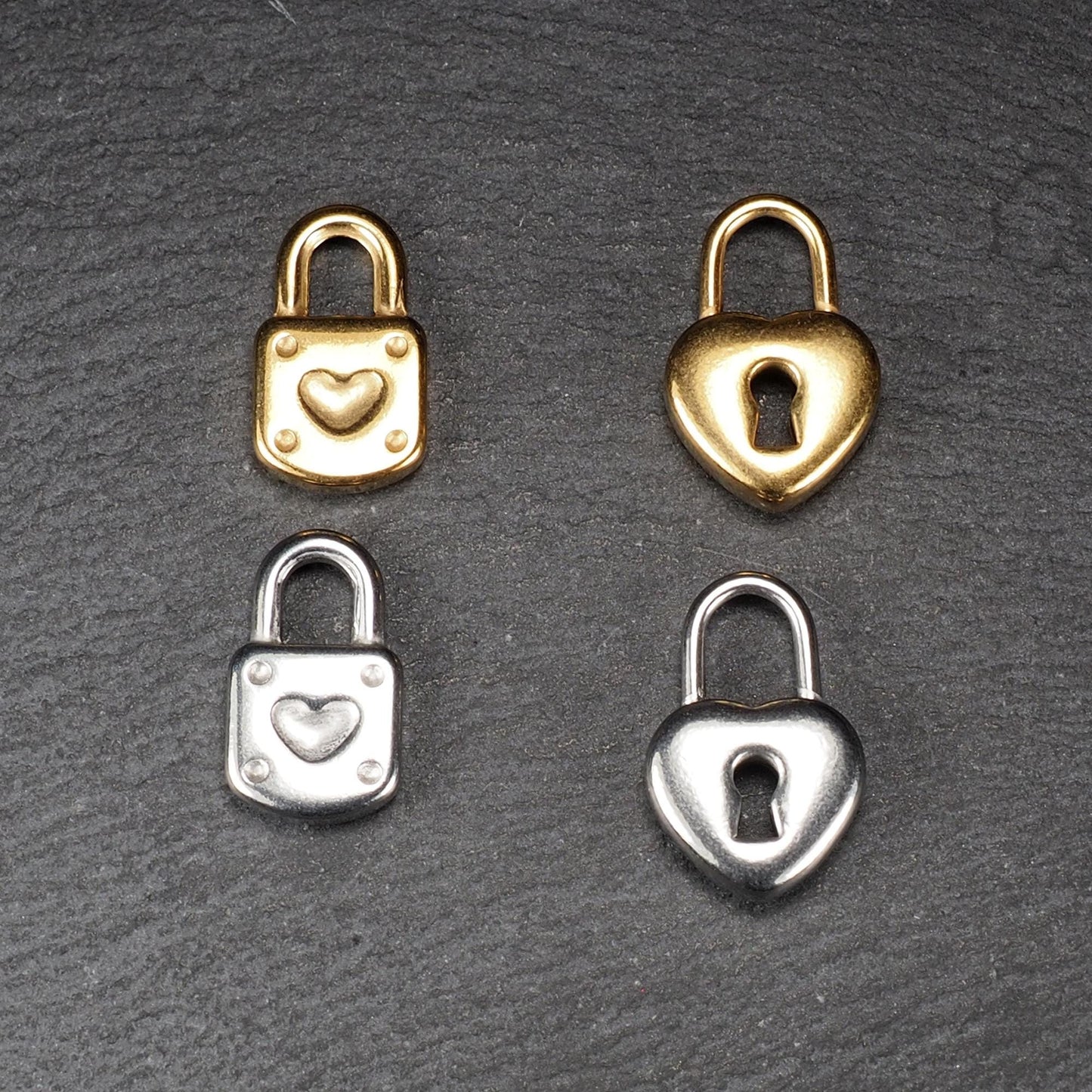 Vier glänzende Metallanhänger auf dunklem Untergrund: zwei vergoldete und zwei silberne Schloss- und Herz-Schlüssel-Motiv-Anhänger mit sichtbarer Öse.