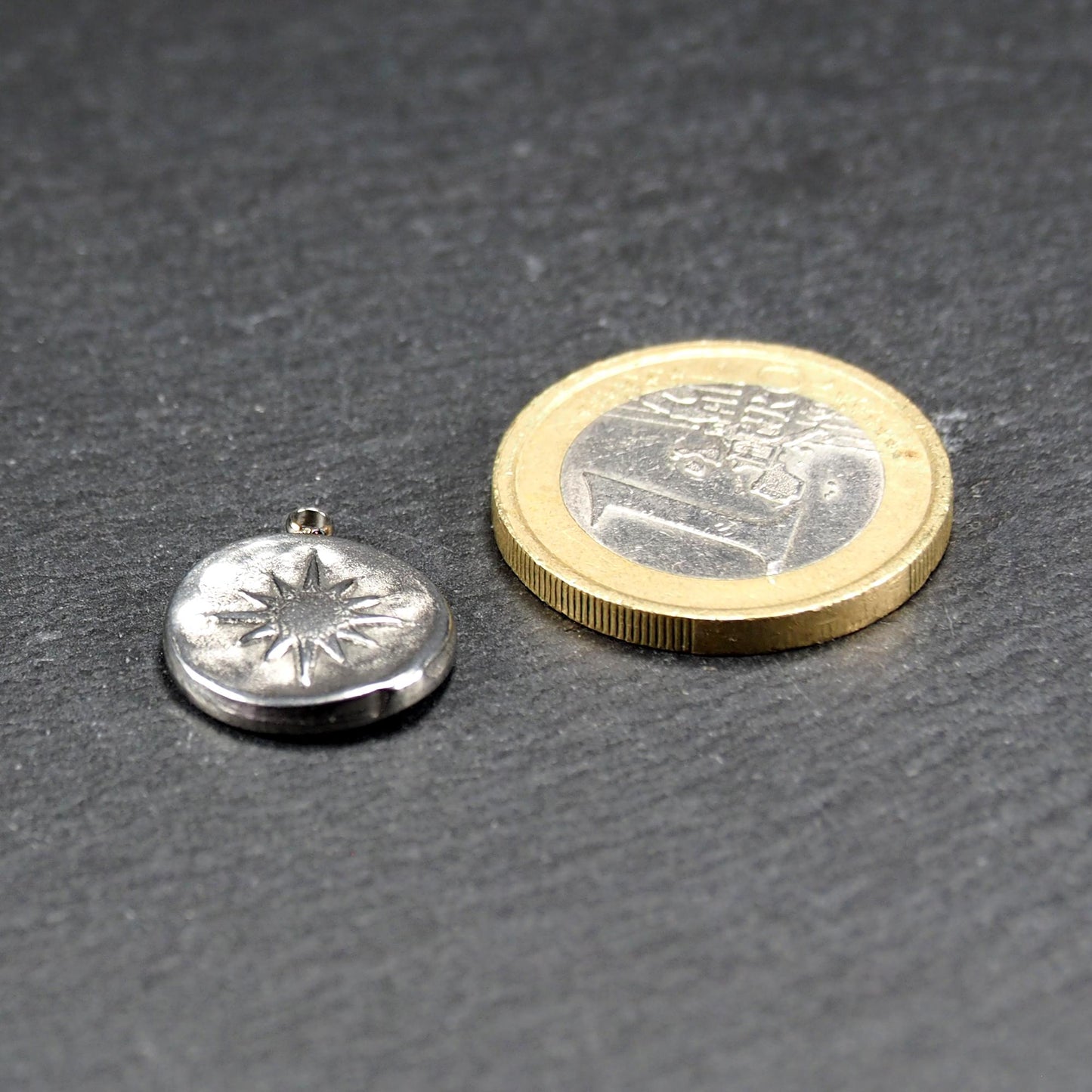 Runder silberner Metallanhänger mit reliefartigem Sonnen- und Kompassmotiv, mit Öse oben; neben einer Ein-Euro-Münze zur Größendarstellung.