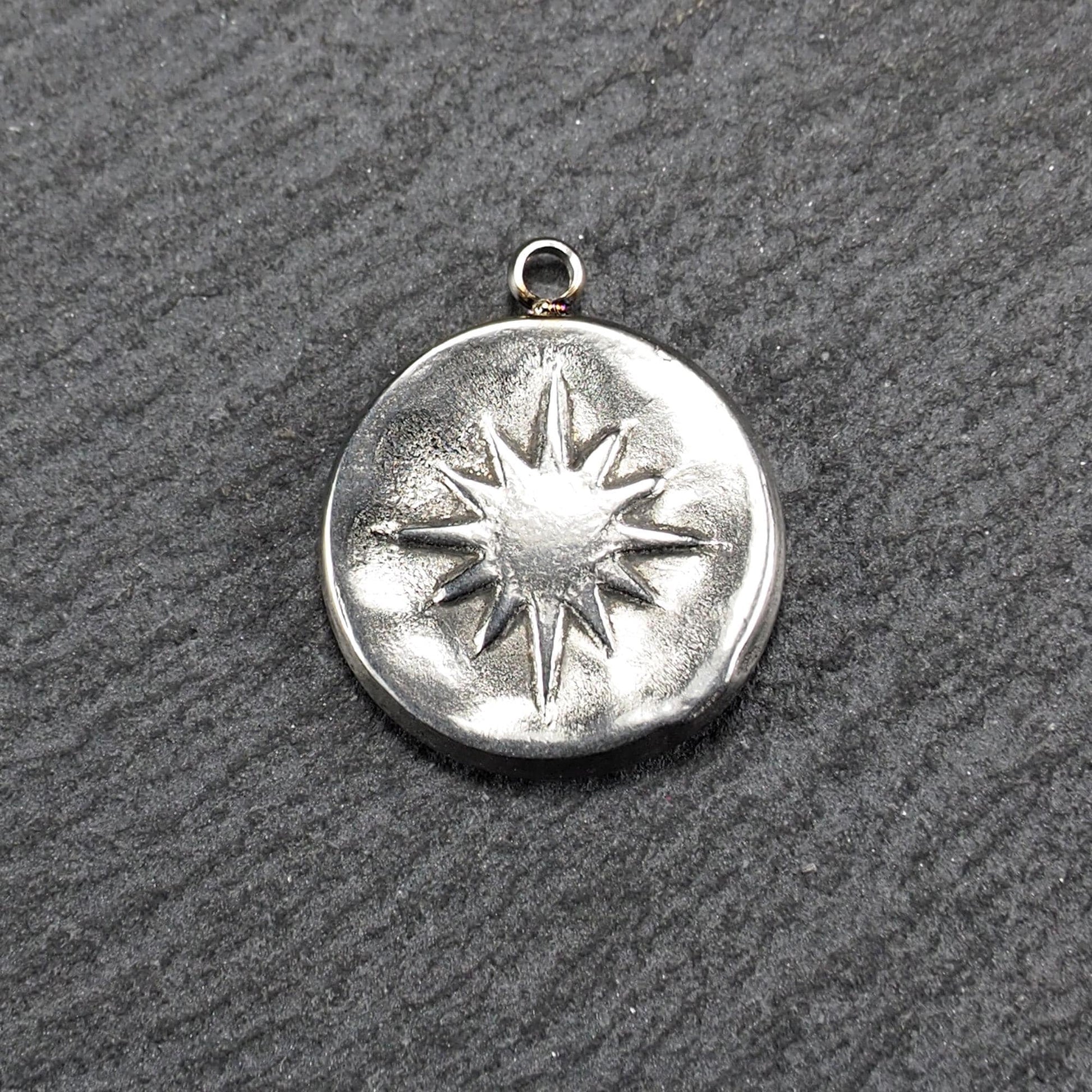Runder silberfarbener Metallanhänger mit leicht strukturierter, glänzender Oberfläche, reliefierter Achtstrahlsonne und kleiner Aufhängeöse oben.