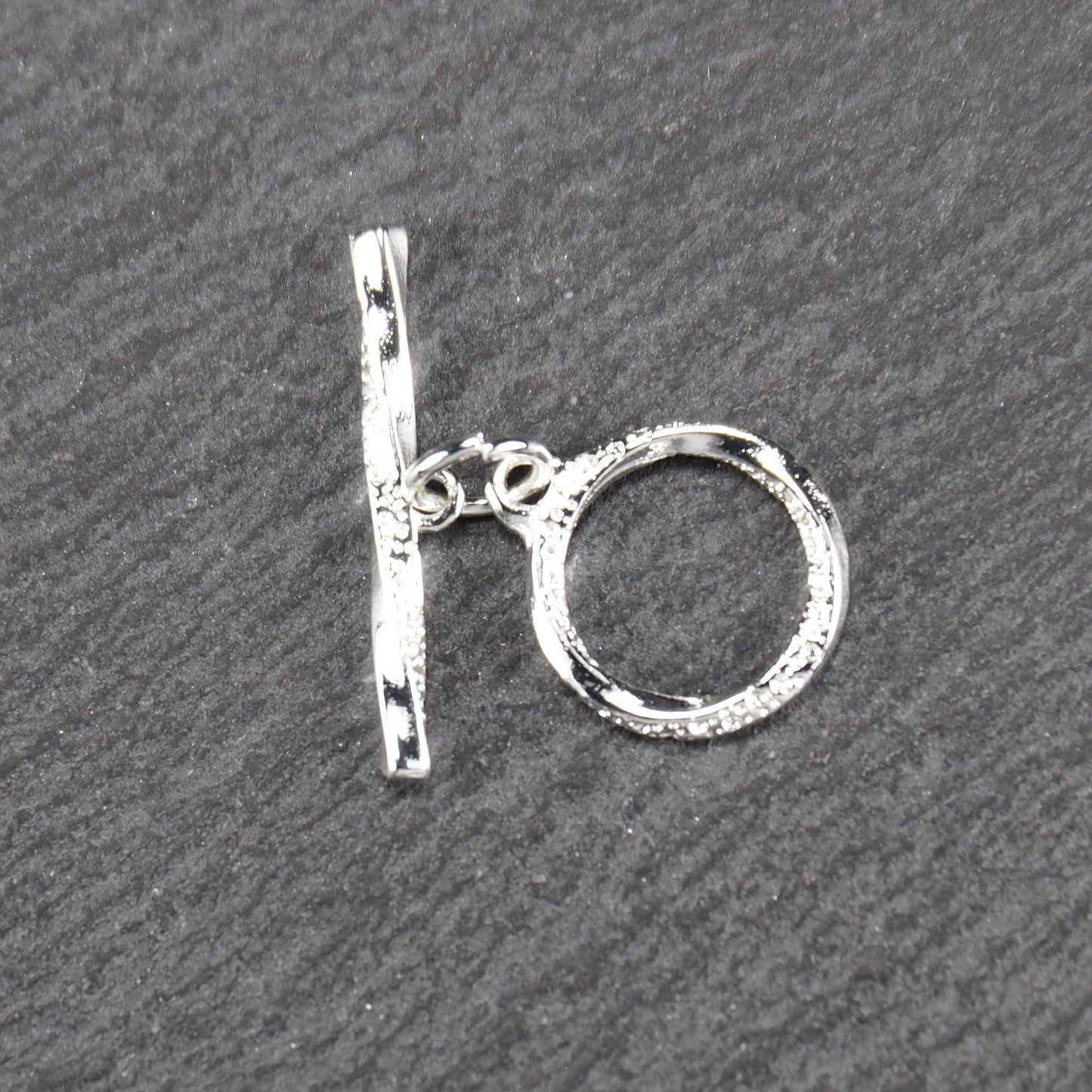 Knebelverschluss (T-Bar) mit silberner, strukturierter Oberfläche, bestehend aus einem horizontalen Stab und einer hängenden, ringförmigen offenen Öse.