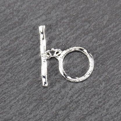 Knebelverschluss (T-Bar) mit silberner, strukturierter Oberfläche, bestehend aus einem horizontalen Stab und einer hängenden, ringförmigen offenen Öse.