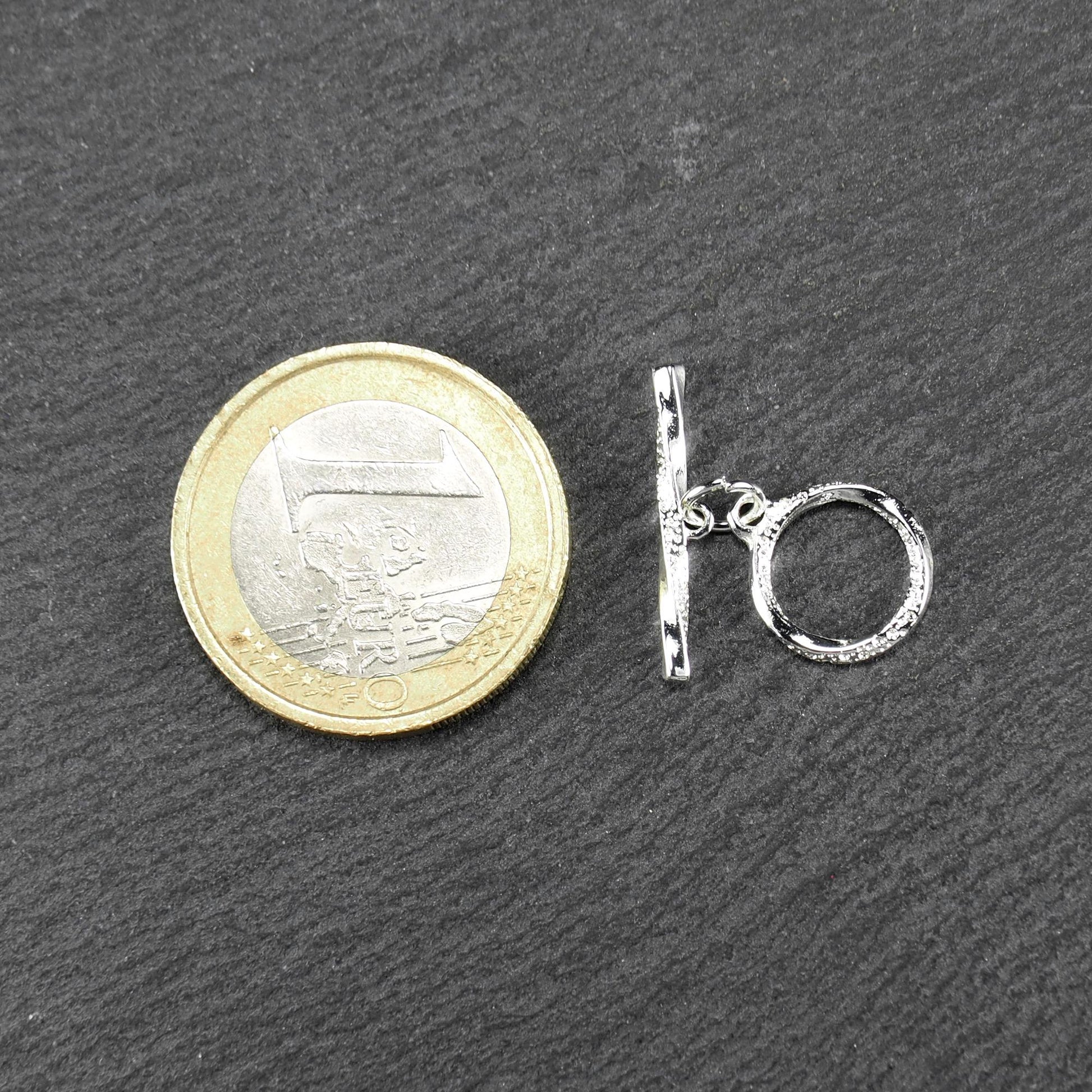 Knebelverschluss (T-Bar) mit silberner, strukturierter Oberfläche, bestehend aus einem horizontalen Stab und einer hängenden, ringförmigen offenen Öse.