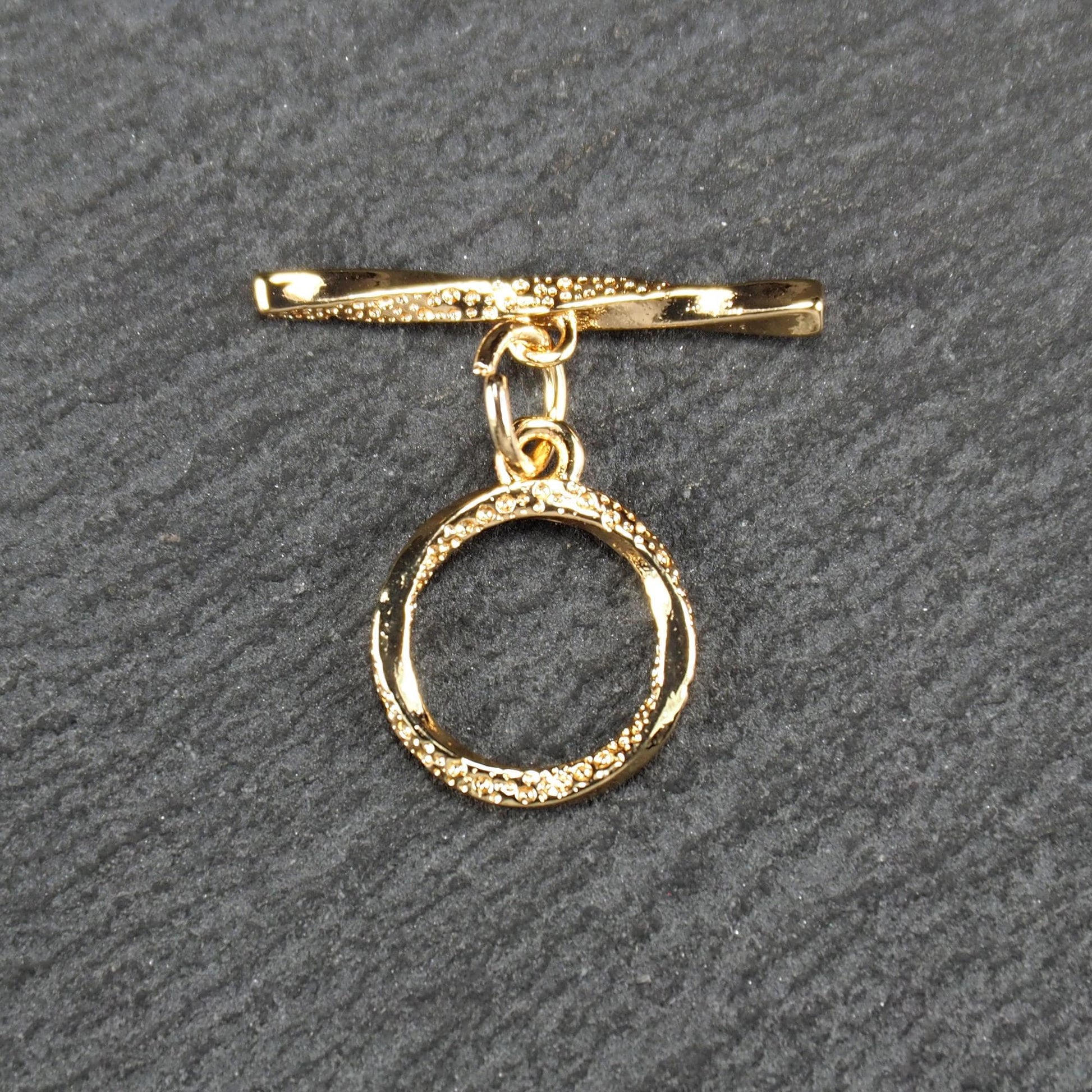 Knebelverschluss, goldfarben, mit runder durchbrochener Öse mit punktierter Struktur und horizontalem Stab, verbunden durch kleine Ringe.