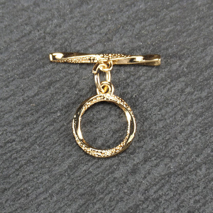 Knebelverschluss, goldfarben, mit runder durchbrochener Öse mit punktierter Struktur und horizontalem Stab, verbunden durch kleine Ringe.