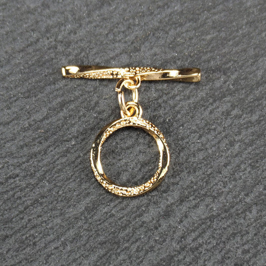 Knebelverschluss, goldfarben, mit runder durchbrochener Öse mit punktierter Struktur und horizontalem Stab, verbunden durch kleine Ringe.