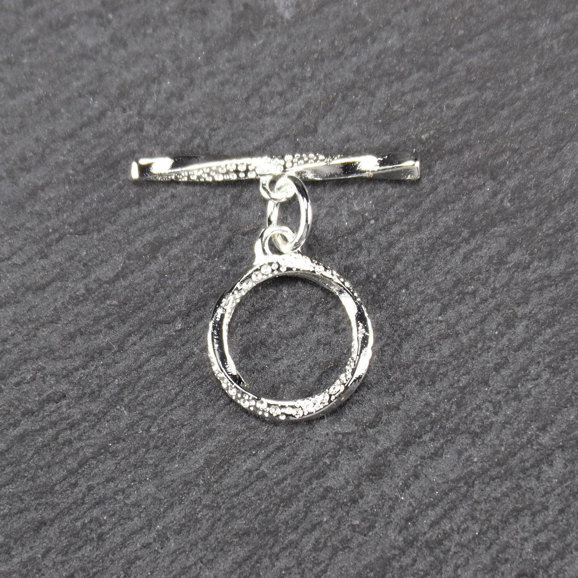 Knebelverschluss (T-Bar) mit silberner, strukturierter Oberfläche, bestehend aus einem horizontalen Stab und einer hängenden, ringförmigen offenen Öse.