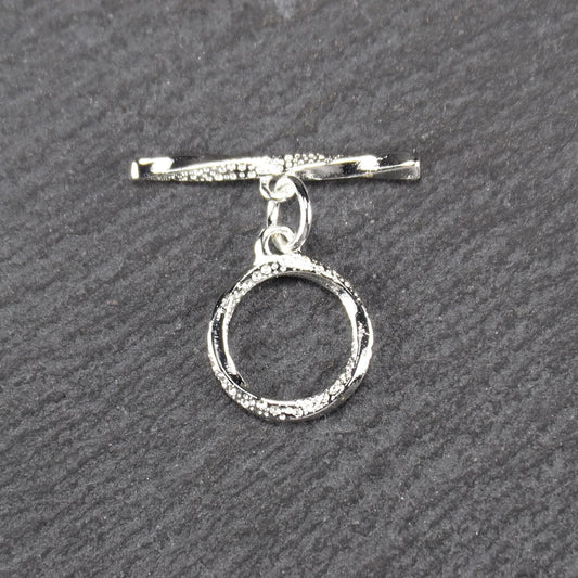 Knebelverschluss (T-Bar) mit silberner, strukturierter Oberfläche, bestehend aus einem horizontalen Stab und einer hängenden, ringförmigen offenen Öse.