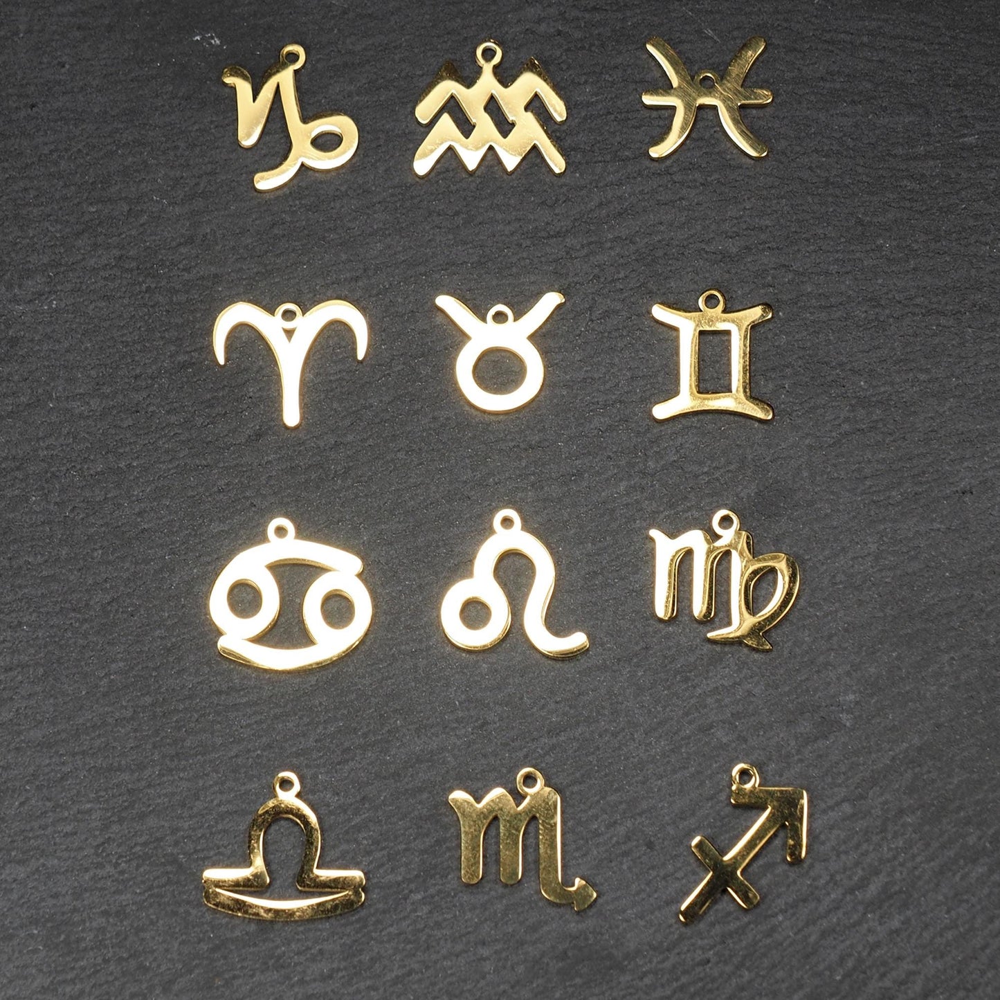 Zwölf flache, vergoldete Sternzeichen-Anhänger aus Metall mit minimalistischer, durchbrochener Kontur und integrierter Öse auf dunklem Hintergrund.