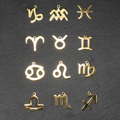 Zwölf flache, vergoldete Sternzeichen-Anhänger aus Metall mit minimalistischer, durchbrochener Kontur und integrierter Öse auf dunklem Hintergrund.