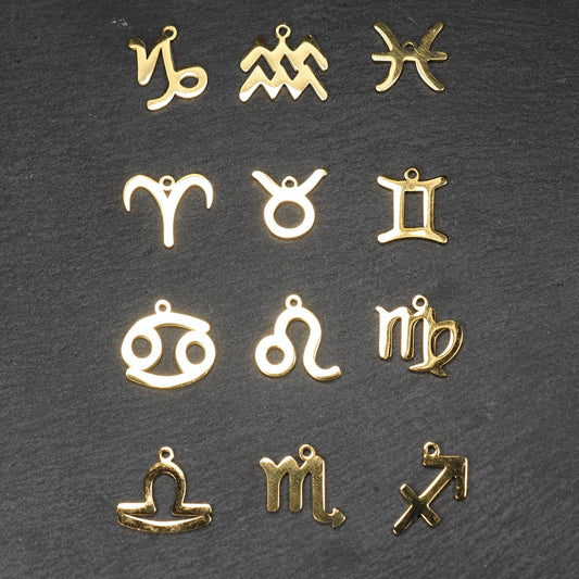 Zwölf flache, vergoldete Sternzeichen-Anhänger aus Metall mit minimalistischer, durchbrochener Kontur und integrierter Öse auf dunklem Hintergrund.