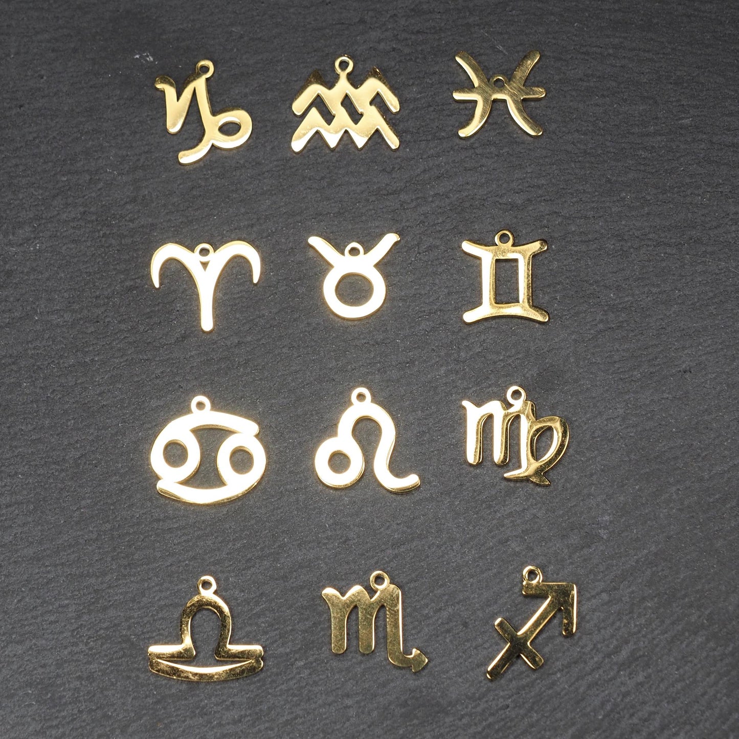 Zwölf flache, vergoldete Sternzeichen-Anhänger aus Metall mit minimalistischer, durchbrochener Kontur und integrierter Öse auf dunklem Hintergrund.