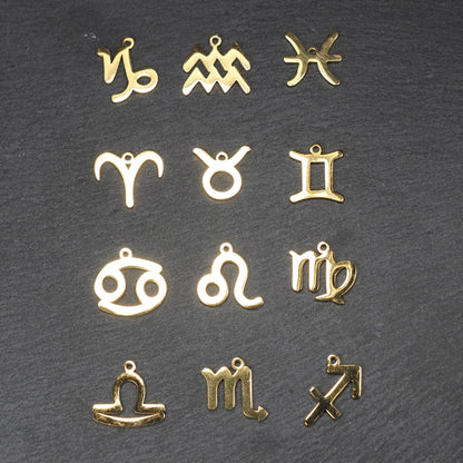 Zwölf flache, vergoldete Sternzeichen-Anhänger aus Metall mit minimalistischer, durchbrochener Kontur und integrierter Öse auf dunklem Hintergrund.
