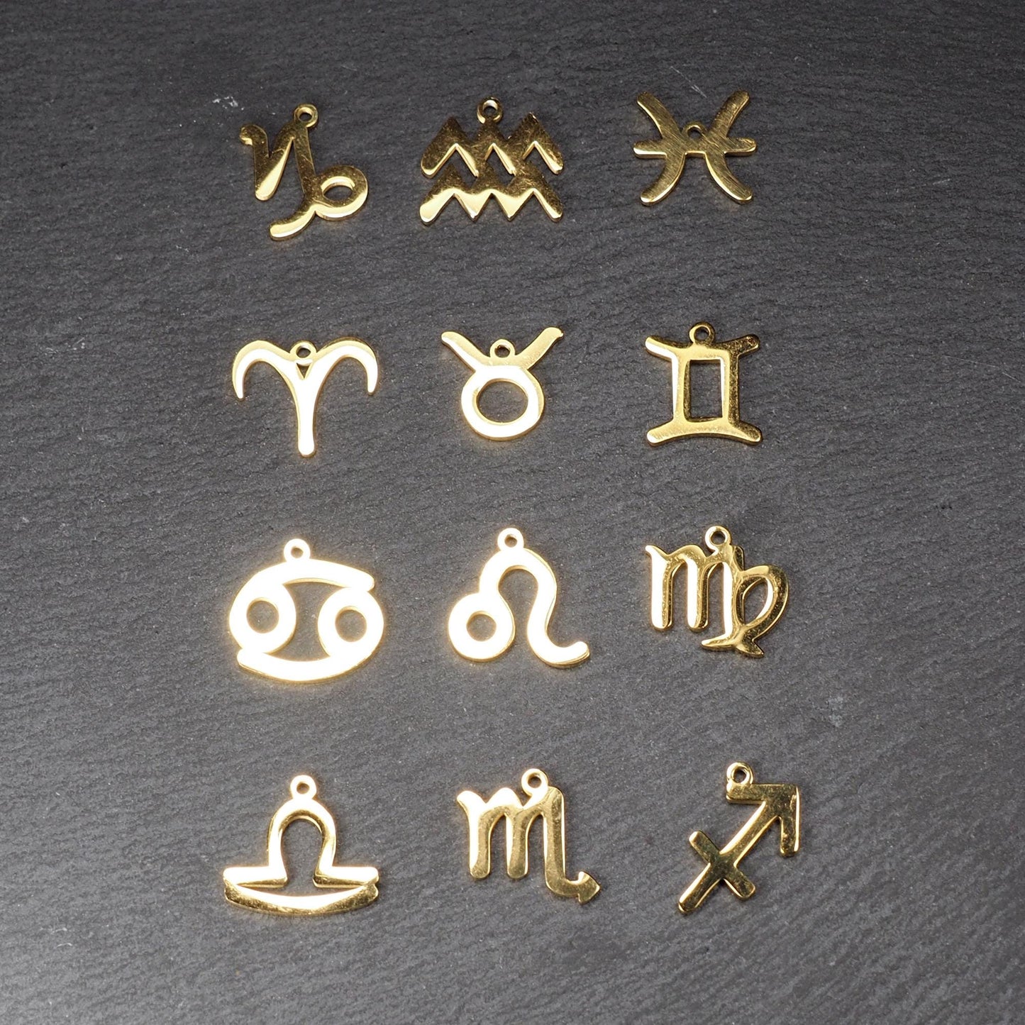 Zwölf flache, vergoldete Sternzeichen-Anhänger aus Metall mit minimalistischer, durchbrochener Kontur und integrierter Öse auf dunklem Hintergrund.
