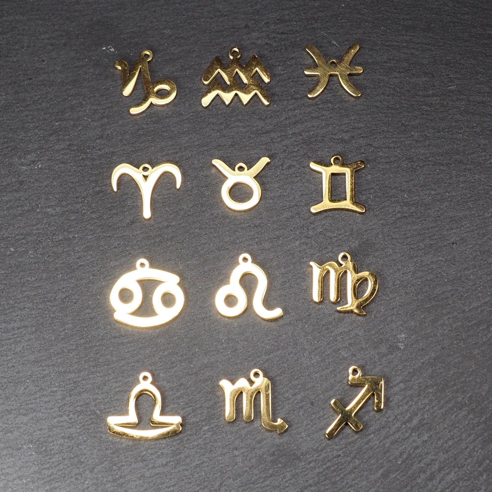 Zwölf flache, vergoldete Sternzeichen-Anhänger aus Metall mit minimalistischer, durchbrochener Kontur und integrierter Öse auf dunklem Hintergrund.