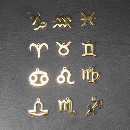 Zwölf flache, vergoldete Sternzeichen-Anhänger aus Metall mit minimalistischer, durchbrochener Kontur und integrierter Öse auf dunklem Hintergrund.