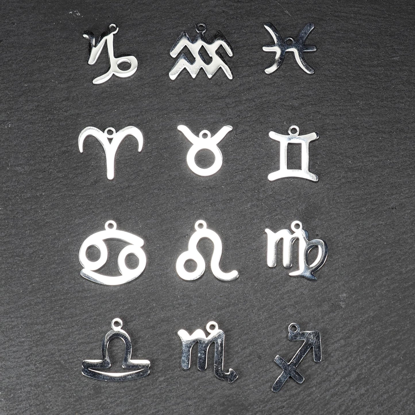 Zwölf flache silberfarbene Edelstahl-Anhänger als durchbrochene Sternzeichensymbole mit glänzender Oberfläche und je einer Öse auf dunklem Hintergrund.