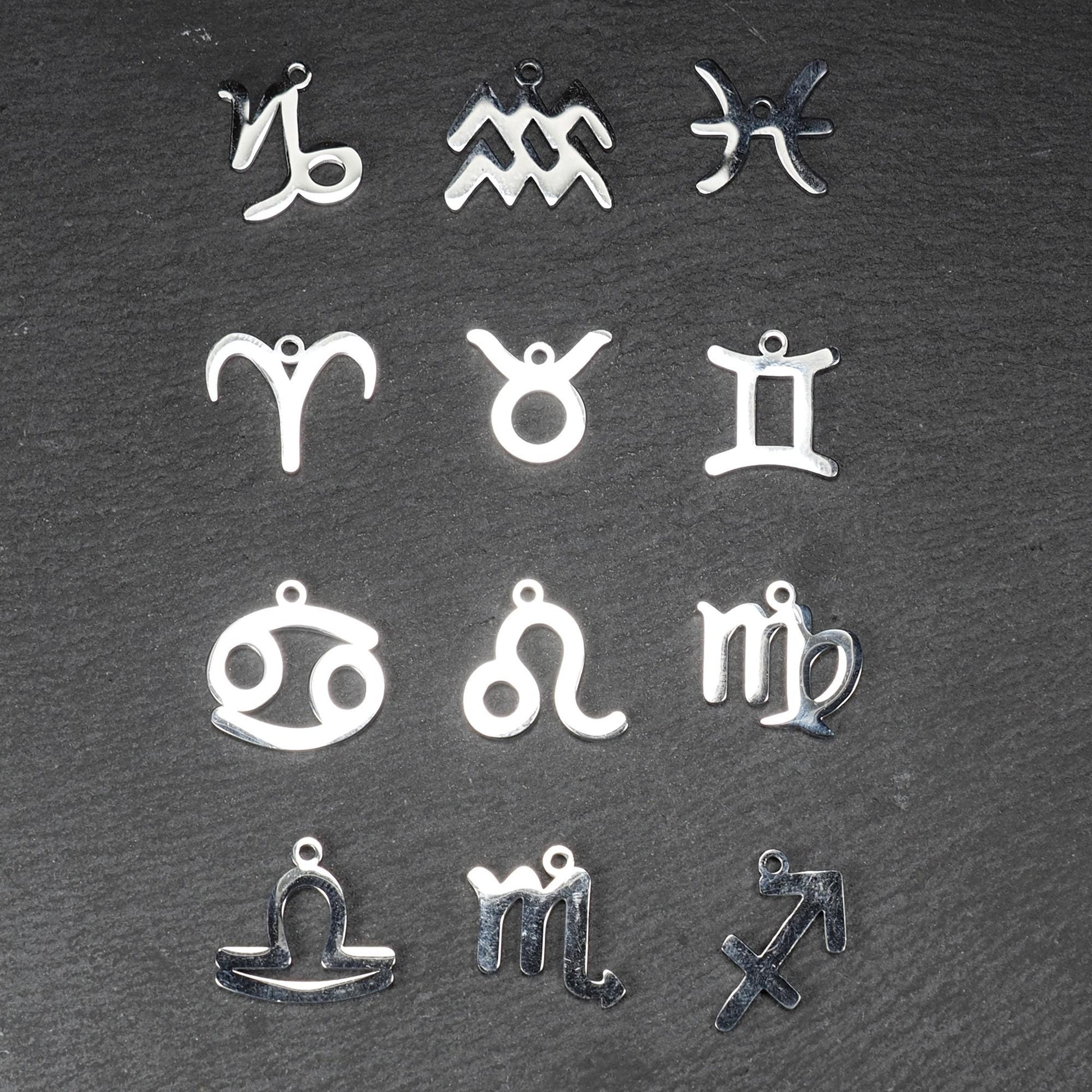Zwölf flache silberfarbene Edelstahl-Anhänger als durchbrochene Sternzeichensymbole mit glänzender Oberfläche und je einer Öse auf dunklem Hintergrund.