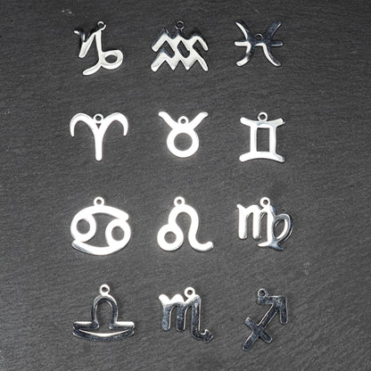 Zwölf flache silberfarbene Edelstahl-Anhänger als durchbrochene Sternzeichensymbole mit glänzender Oberfläche und je einer Öse auf dunklem Hintergrund.
