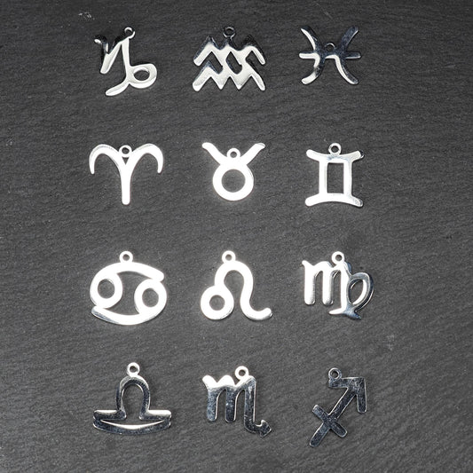 Zwölf flache silberfarbene Edelstahl-Anhänger als durchbrochene Sternzeichensymbole mit glänzender Oberfläche und je einer Öse auf dunklem Hintergrund.