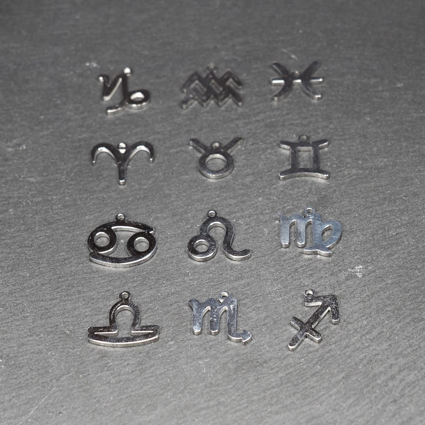 Zwölf flache silberfarbene Edelstahl-Anhänger als durchbrochene Sternzeichensymbole mit glänzender Oberfläche und je einer Öse auf dunklem Hintergrund.