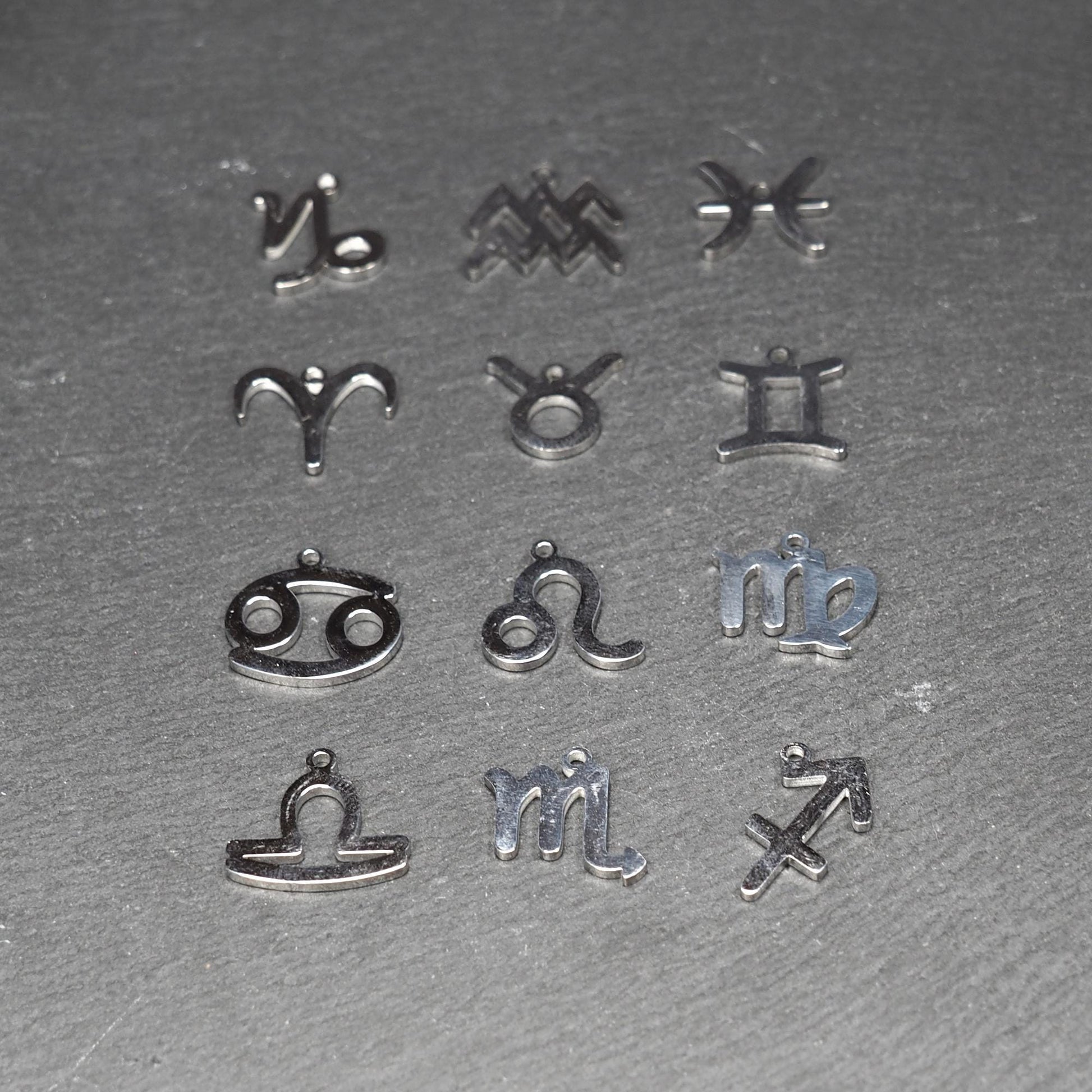 Zwölf flache silberfarbene Edelstahl-Anhänger als durchbrochene Sternzeichensymbole mit glänzender Oberfläche und je einer Öse auf dunklem Hintergrund.