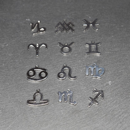 Zwölf flache silberfarbene Edelstahl-Anhänger als durchbrochene Sternzeichensymbole mit glänzender Oberfläche und je einer Öse auf dunklem Hintergrund.
