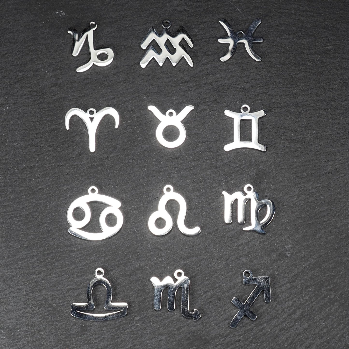 Zwölf flache silberfarbene Edelstahl-Anhänger als durchbrochene Sternzeichensymbole mit glänzender Oberfläche und je einer Öse auf dunklem Hintergrund.