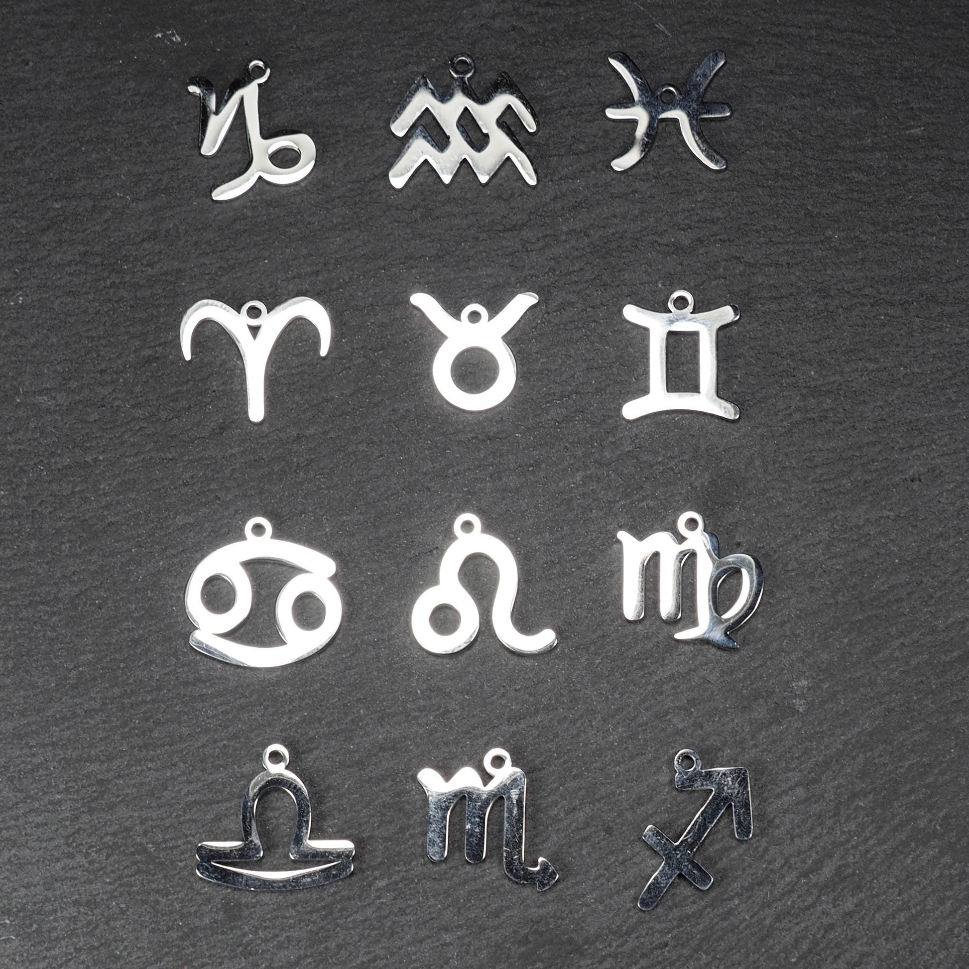Zwölf flache silberfarbene Edelstahl-Anhänger als durchbrochene Sternzeichensymbole mit glänzender Oberfläche und je einer Öse auf dunklem Hintergrund.