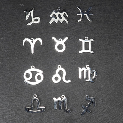 Zwölf flache silberfarbene Edelstahl-Anhänger als durchbrochene Sternzeichensymbole mit glänzender Oberfläche und je einer Öse auf dunklem Hintergrund.
