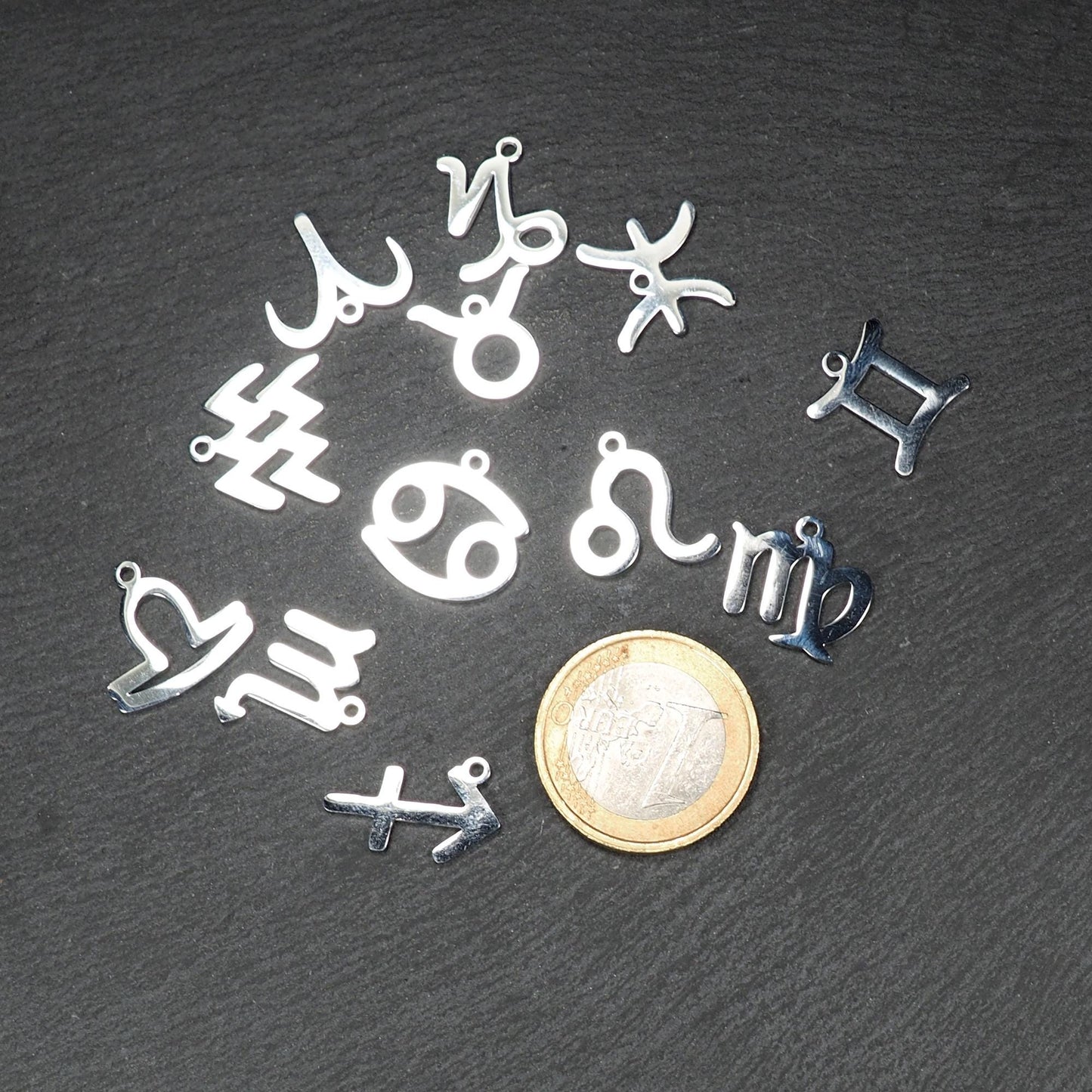 Mehrere flache silberne Sternzeichen-Anhänger aus glänzendem Metall mit Öse, neben Ein-Euro-Münze als Größenvergleich.