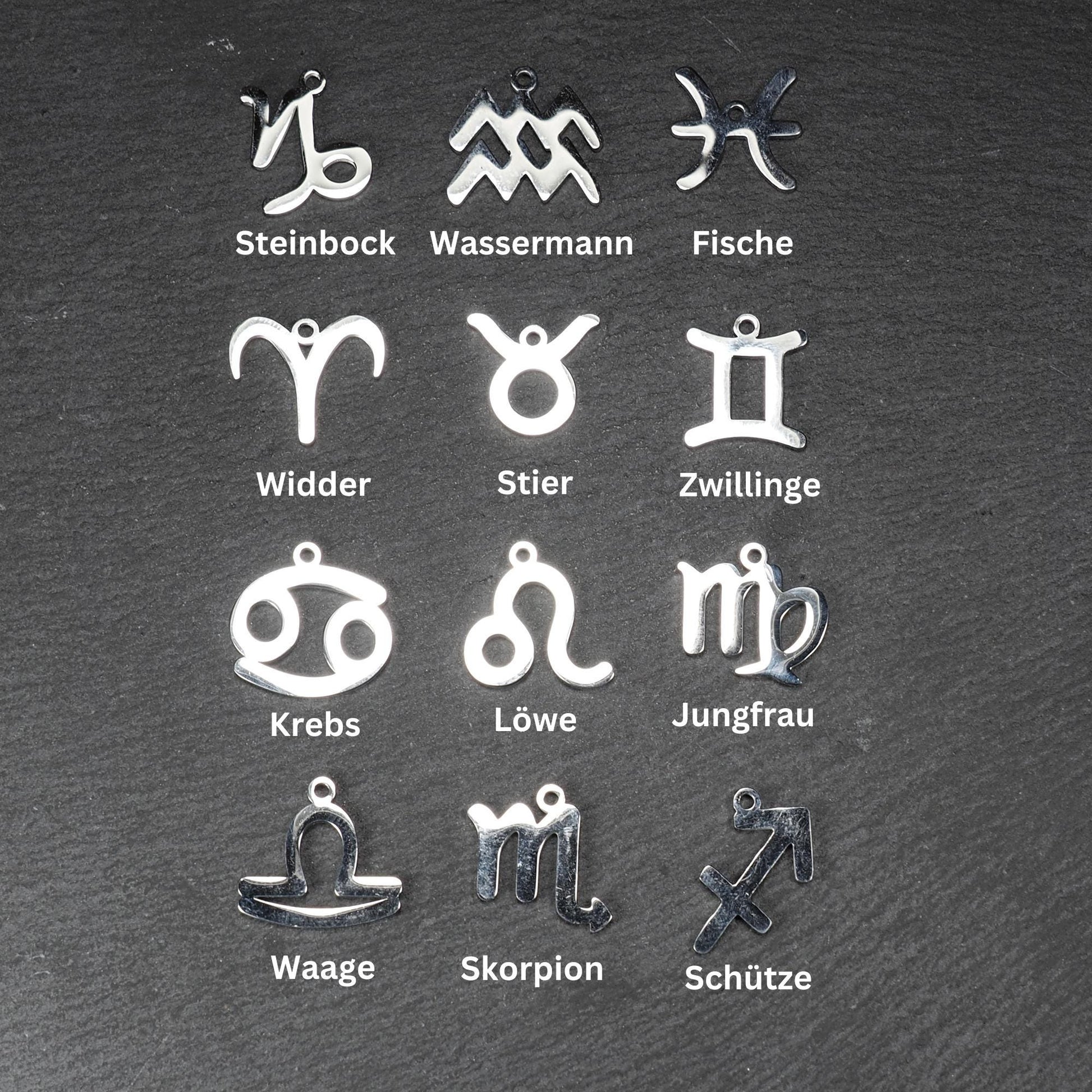 Zwölf flache silberfarbene Edelstahl-Anhänger als durchbrochene Sternzeichensymbole mit glänzender Oberfläche und je einer Öse auf dunklem Hintergrund.