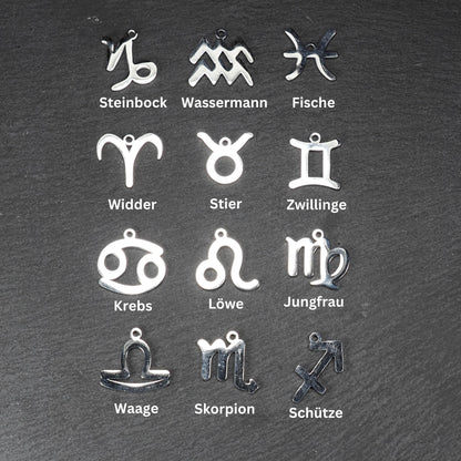Zwölf flache silberfarbene Edelstahl-Anhänger als durchbrochene Sternzeichensymbole mit glänzender Oberfläche und je einer Öse auf dunklem Hintergrund.